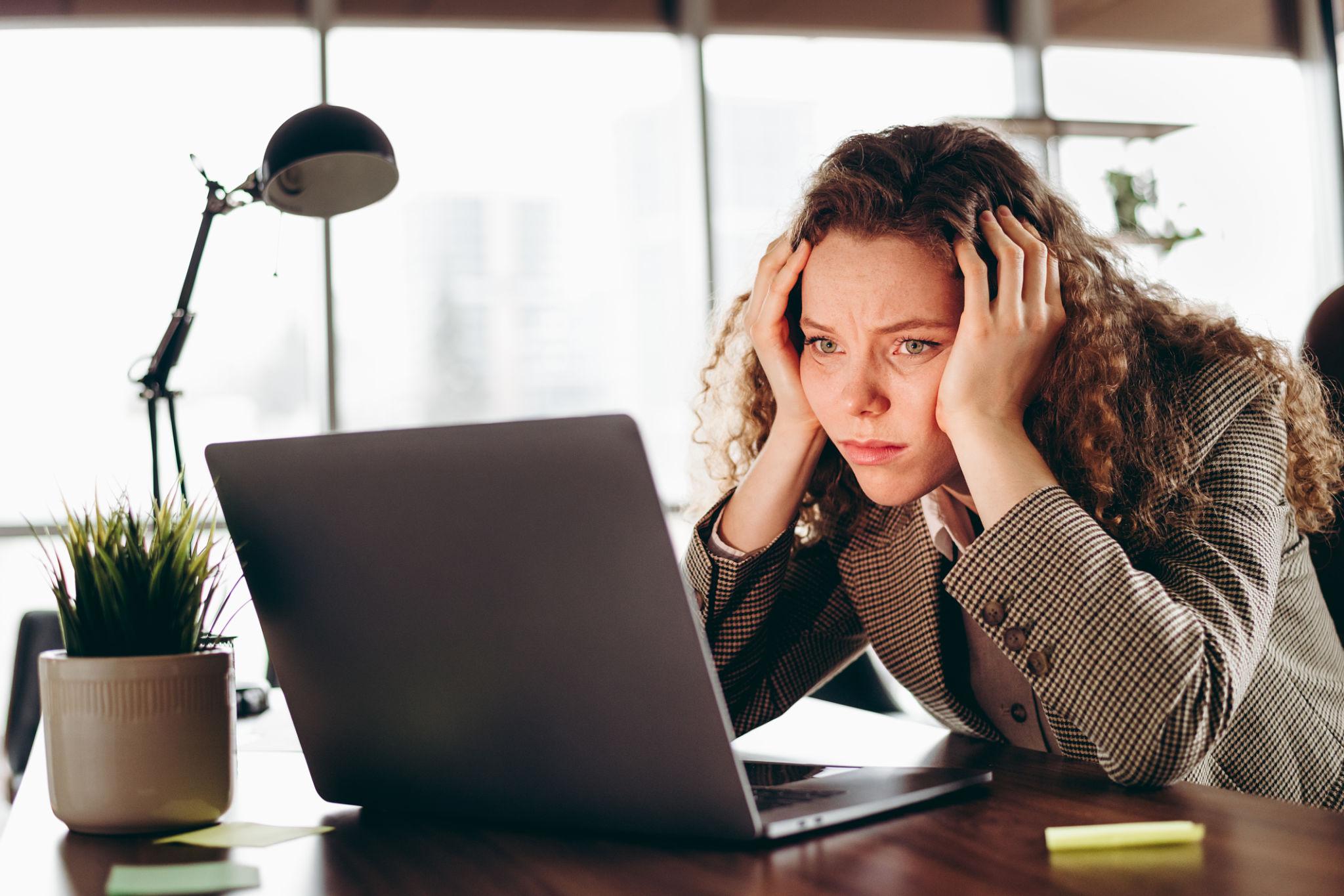 Stressed professional woman holding her head while sitting at a laptop, feeling overwhelmed by workplace anxiety and the pressure to constantly check work messages.